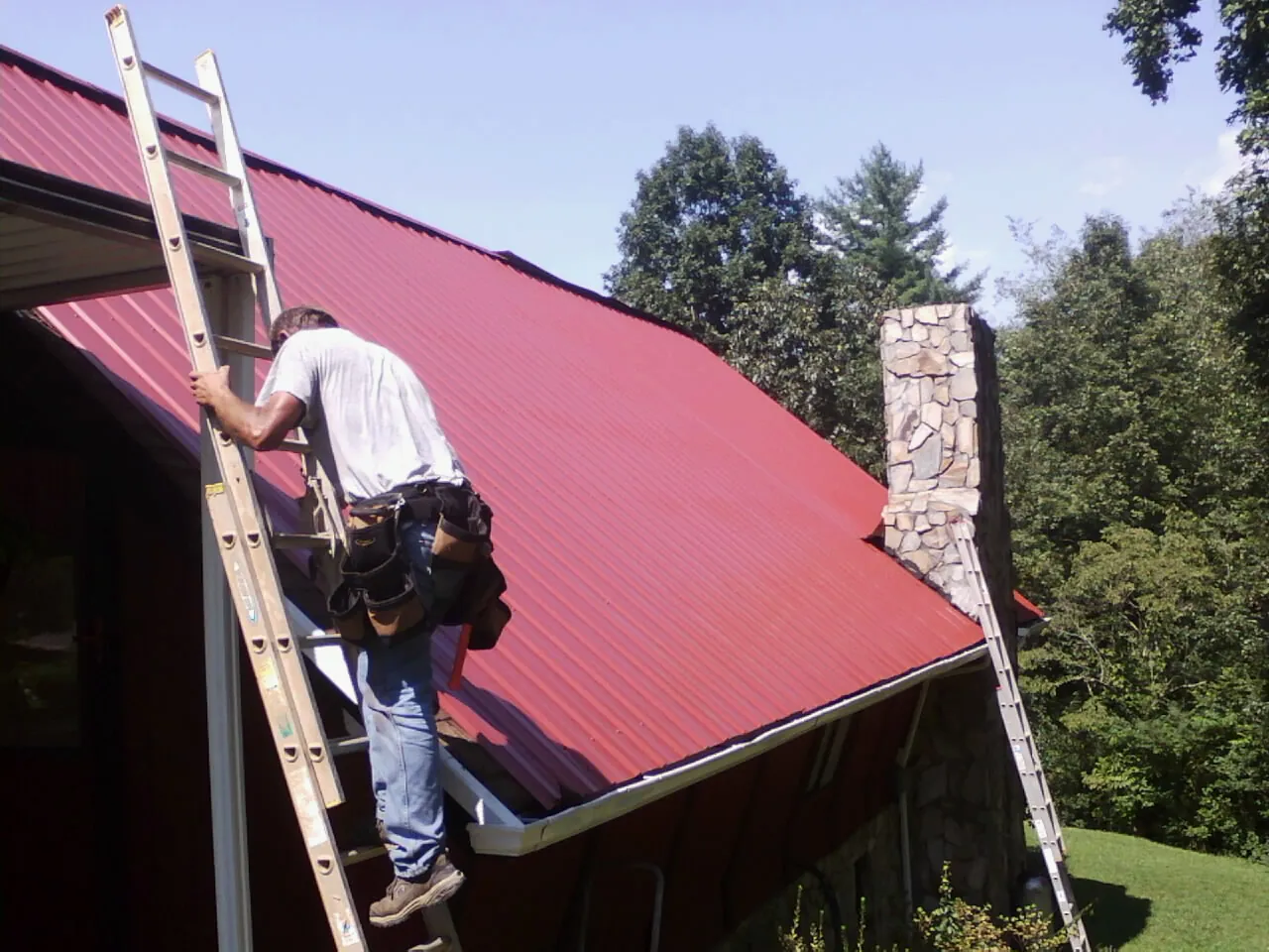 Roofer on a ladder beside a red metal roof and stone chimney — Soffit & Fascia Repair by Roofing Brothers in Lapwai